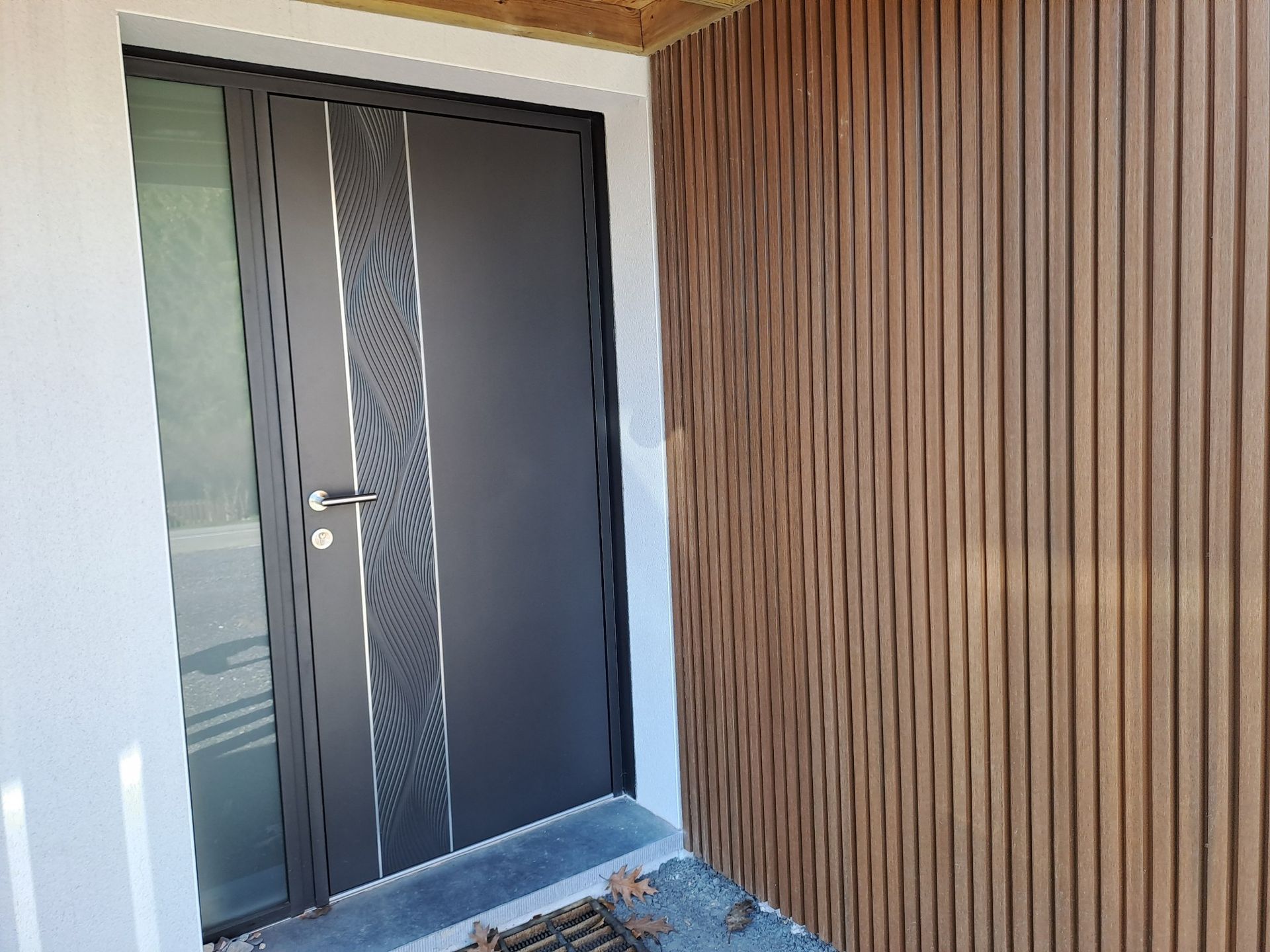 Porte d'entrée gris foncé avec panneau décoratif, à côté d'un mur en lattes de bois marron.