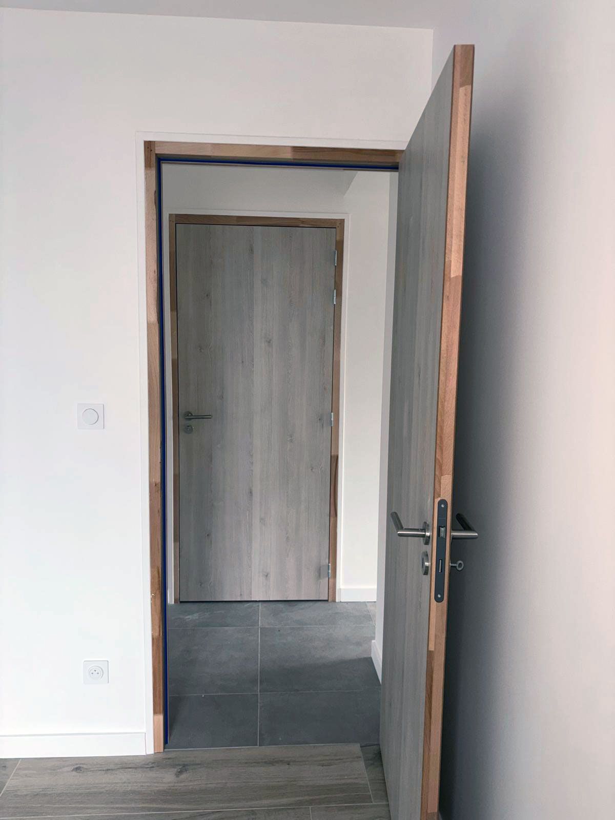 Une porte grise ouverte dans une pièce blanche, révélant une autre porte fermée et un sol en carrelage sombre.