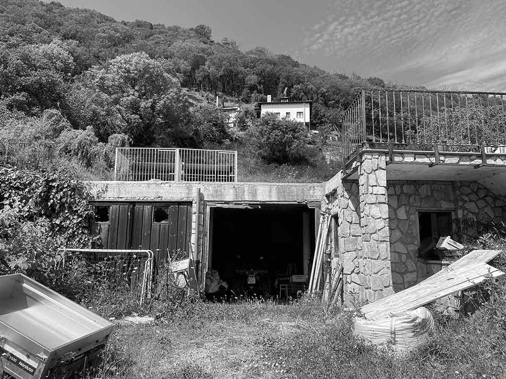 Una estructura en la ladera de una colina con dos garajes abiertos, una fachada de muro de piedra, una terraza superior y una densa vegetación al fondo.