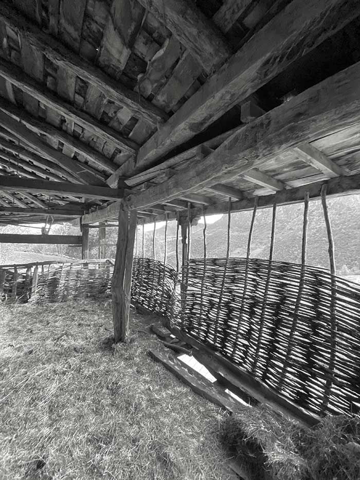 Una estructura rústica de madera, con los laterales abiertos y una cerca de ramas entrelazadas bajo un techo de madera desgastada.