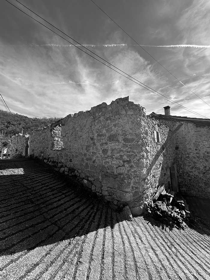 Una imagen en blanco y negro de un muro de piedra rústica que proyecta largas y dramáticas sombras sobre un suelo texturizado bajo un cielo nublado.