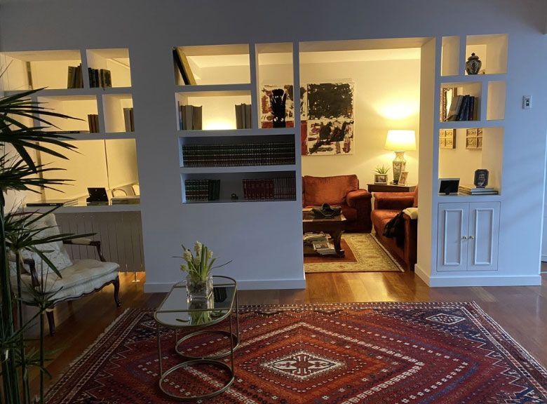 Un luminoso salón cuenta con estanterías blancas empotradas, una alfombra roja estampada, una mesa de centro de cristal y una acogedora zona de estar.