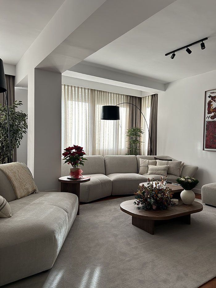 Un salón moderno con sofás curvos de color crema, una mesa de centro redonda de madera, lámparas de pie y cortinas que filtran la luz.