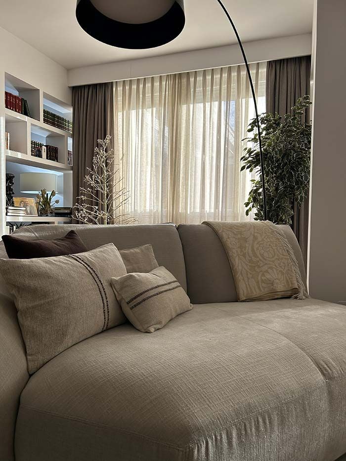 Una acogedora sala de estar con un sofá de tonos neutros, cojines decorativos, una lámpara de pie y cortinas claras que cubren una ventana.