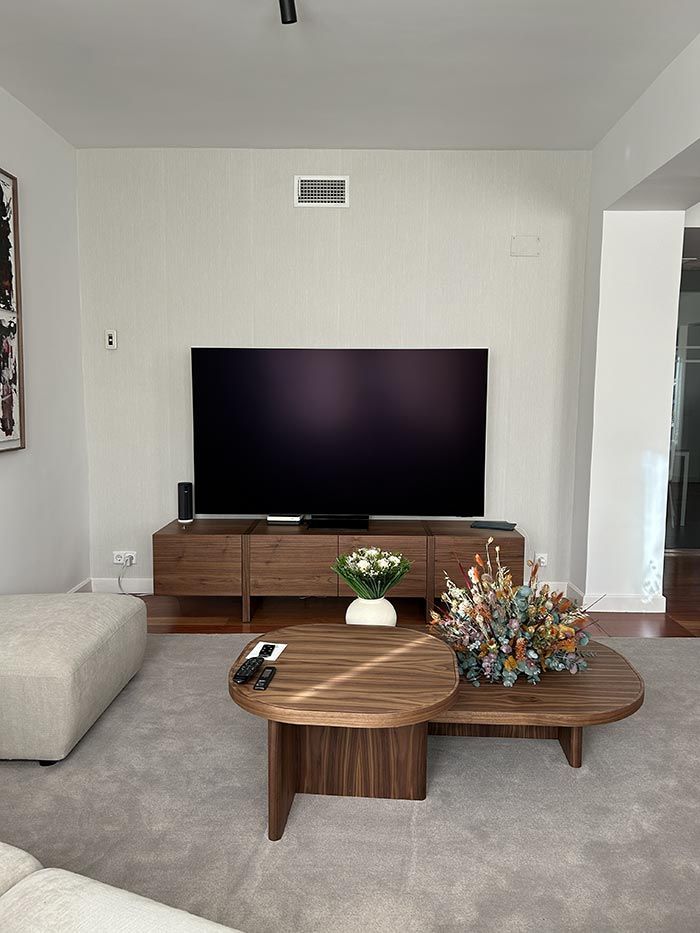 Una sala de estar con un televisor grande sobre un mueble de madera para equipos multimedia, una mesa de centro redonda de madera y una alfombra mullida.