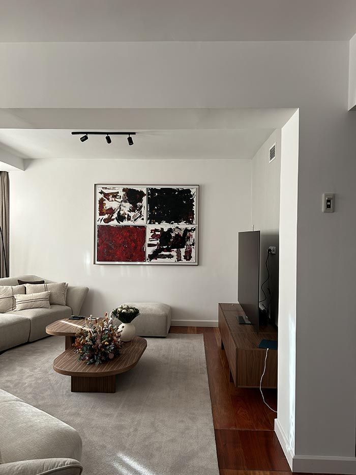 Un salón moderno con un sofá de color neutro, dos mesas de centro de madera, un gran cuadro abstracto y un televisor sobre un soporte.