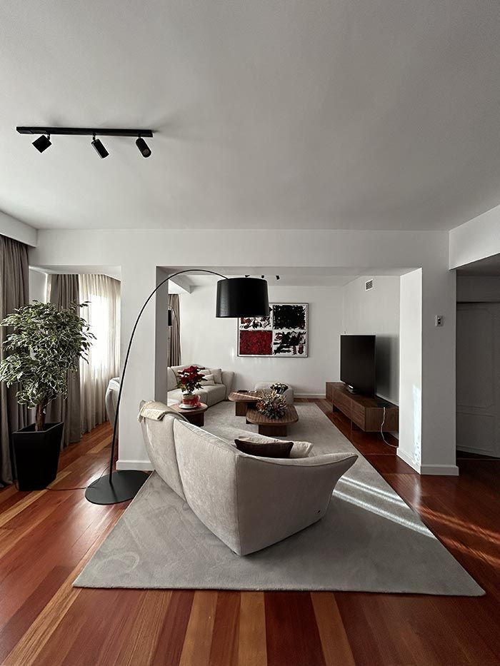 Un salón moderno con un sofá beige sobre una alfombra gris, suelo de madera, una lámpara de pie y un mueble para la televisión con un televisor.