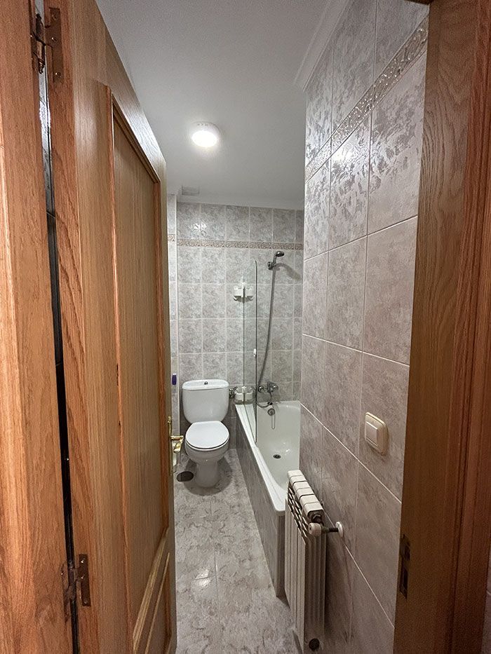 Un baño estrecho, alicatado, con inodoro y bañera, visto a través de una puerta de madera abierta.