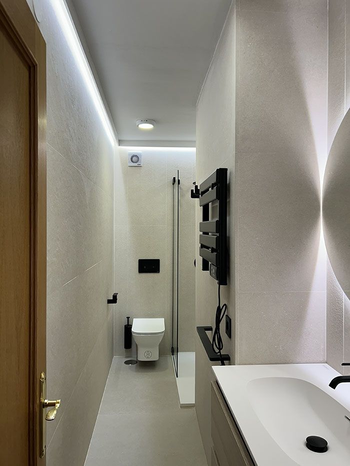 Un baño estrecho y moderno con paredes revestidas de azulejos de color claro, inodoro suspendido, cabina de ducha de cristal y lavabo con tocador.