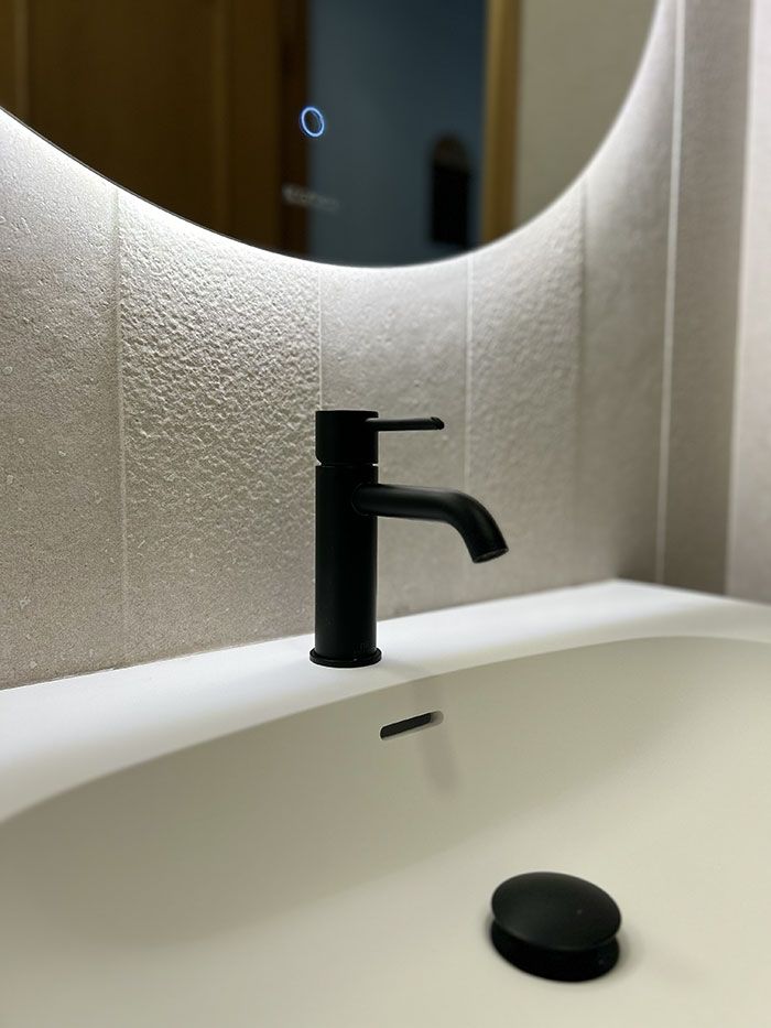 Un lavabo moderno con grifo negro y tapón de desagüe a juego, sobre un panel de protección antisalpicaduras de color claro.