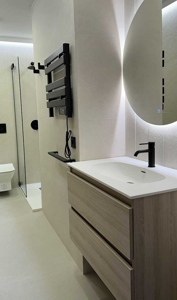 Un baño moderno con tocador con acabado de madera, grifo negro, espejo redondo retroiluminado y ducha a ras de suelo.