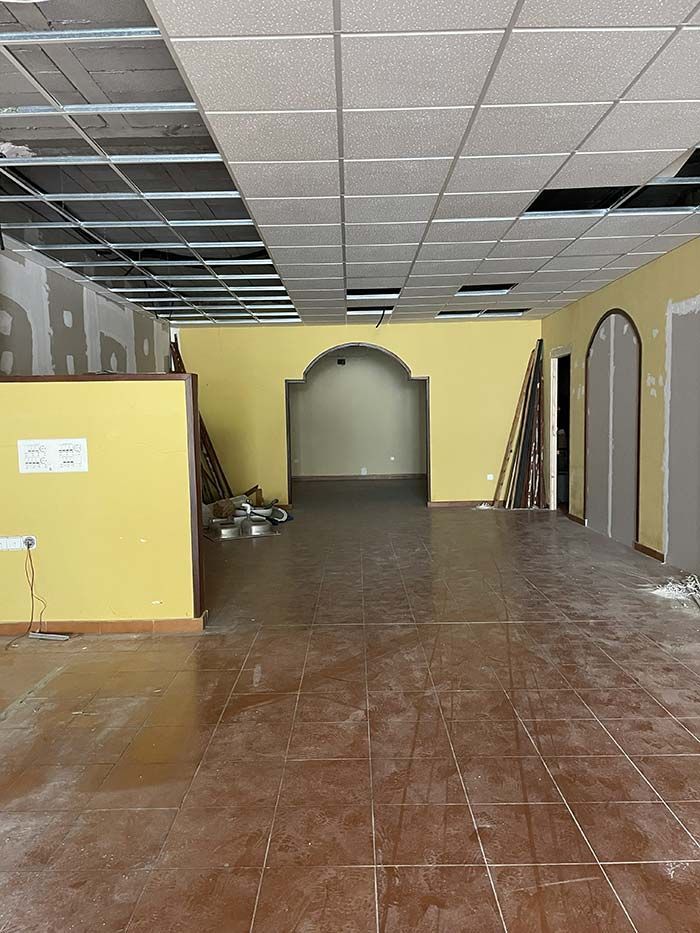 Interior de un local comercial en proceso de renovación con falsos techos sin terminar, paredes amarillas y suelo marrón a la vista.