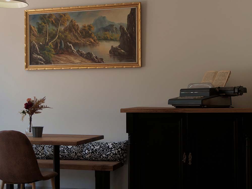 Un mueble de madera oscura con una máquina de escribir encima se encuentra junto a una mesita, un banco y un cuadro de paisaje enmarcado.