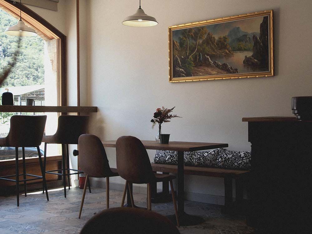 Un acogedor interior de cafetería con una mesa de madera, dos sillas, un banco y un cuadro paisajístico colgado en una pared de color neutro.