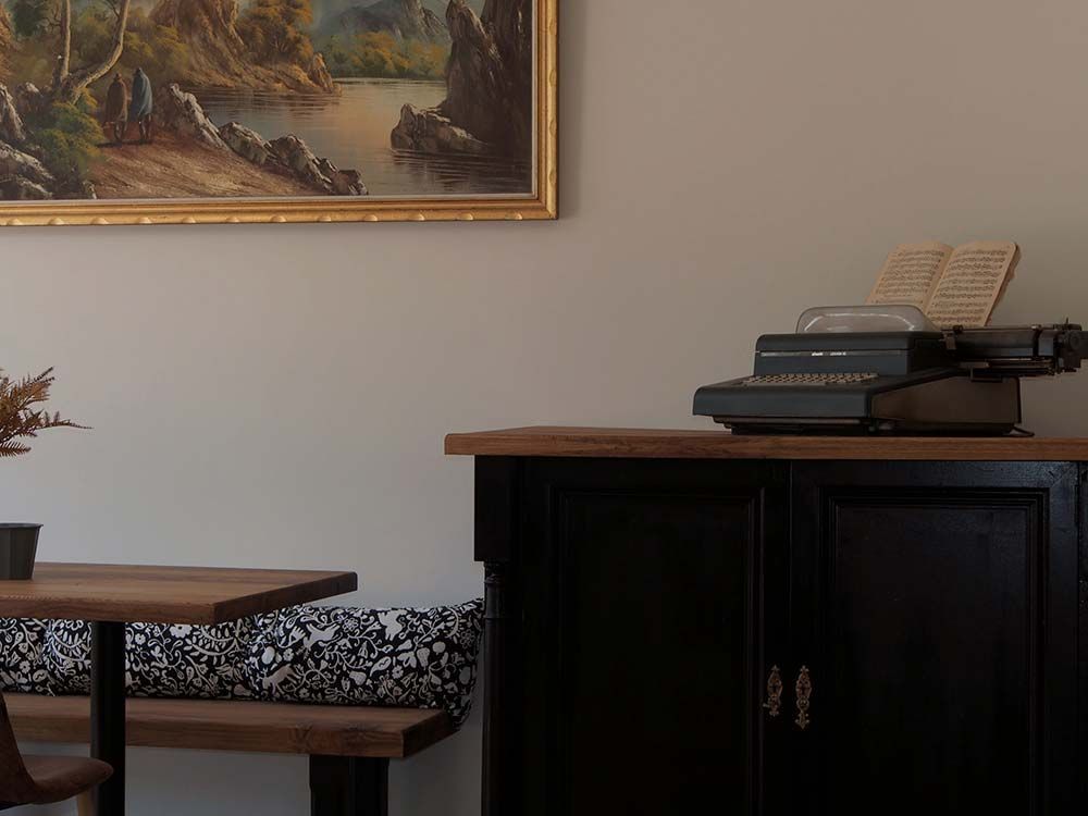 Una máquina de escribir antigua reposa sobre un mueble de madera oscura junto a una mesa de comedor con un banco estampado y un cuadro con un paisaje.