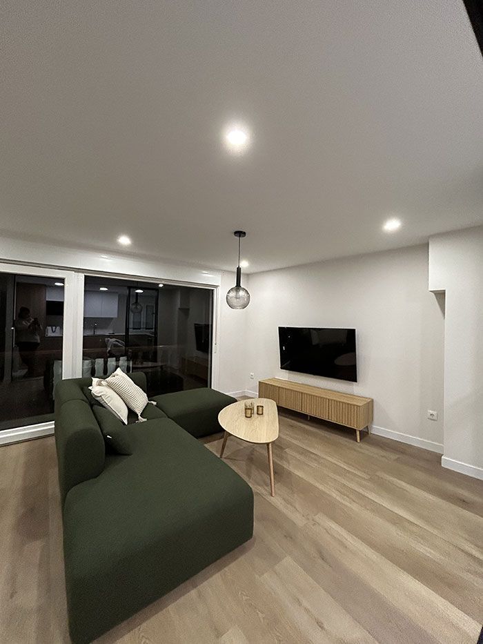 Salón moderno con un gran sofá modular de color verde oscuro, una mesa de centro de madera, un televisor montado en la pared y suelos de madera clara.