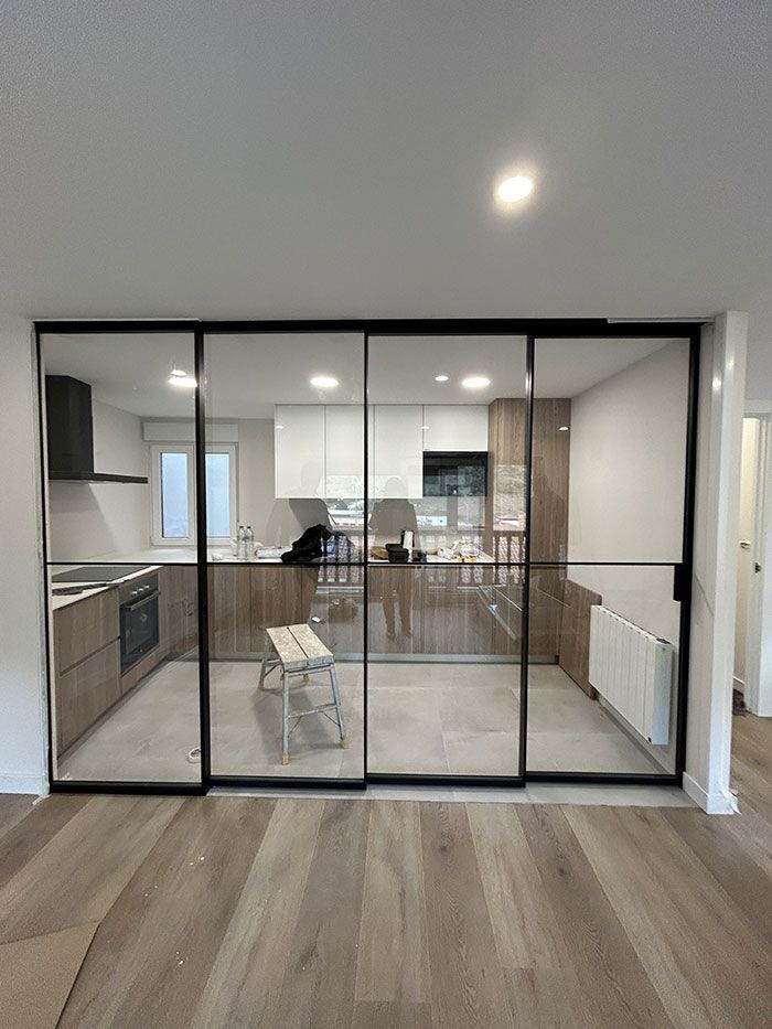 Una cocina moderna vista a través de una mampara corrediza de vidrio de cuatro paneles con marco negro en una sala de estar con piso estilo madera.