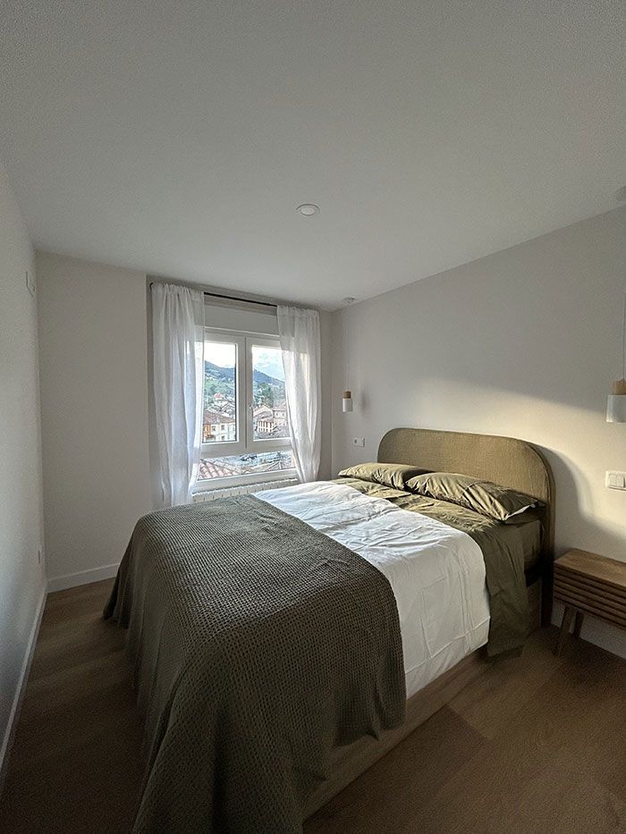 Un dormitorio acogedor con una cama bien hecha, ropa de cama de color verde oliva, cortinas blancas y luz natural que entra por la ventana.