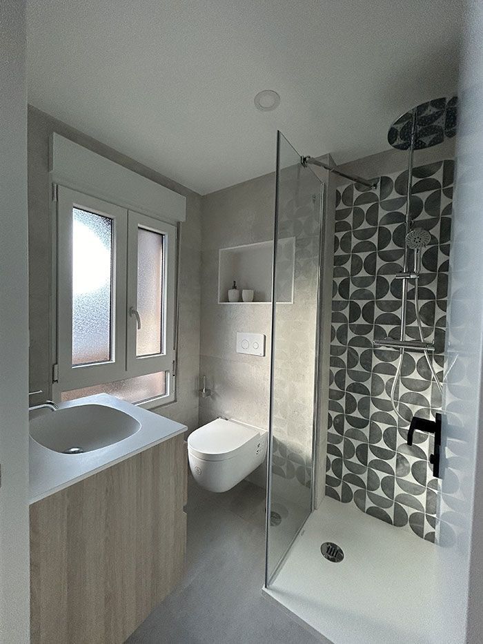 Un baño moderno con tocador de madera clara, inodoro suspendido y ducha a ras de suelo con paredes de azulejos con motivos geométricos.