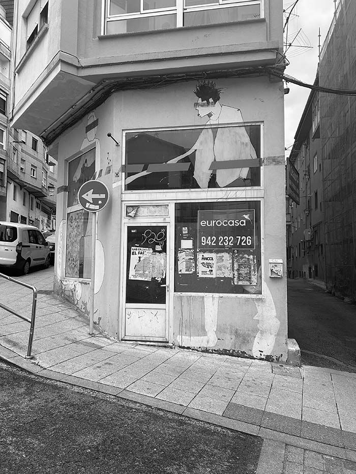 Un edificio en una esquina, de color blanco y negro, con un mural en la pared que representa la parte superior del cuerpo de una persona y carteles en los escaparates.