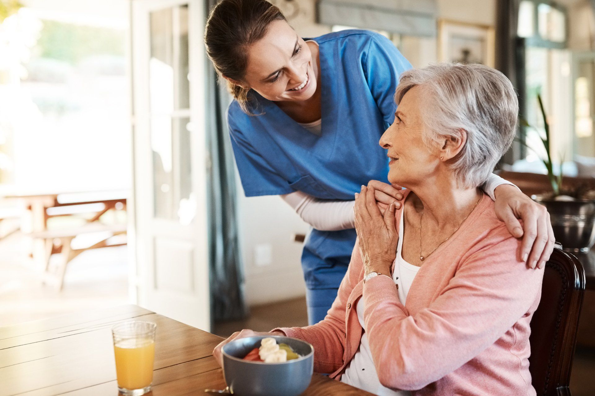 fairpflegt GbR – eine Pflegefachkraft beugt sich lächelnd über eine Seniorin am Esstisch