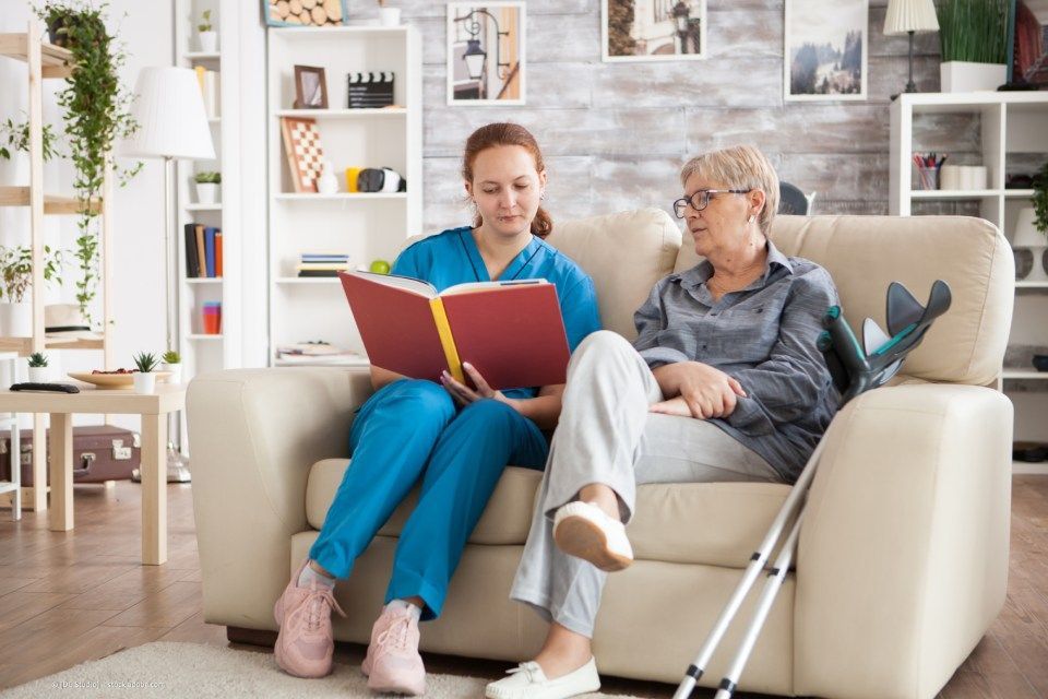 fairpflegt GbR – eine Pflegerin und eine Seniorin mit Krücken sitzen auf dem Sofa und lesen ein Buch