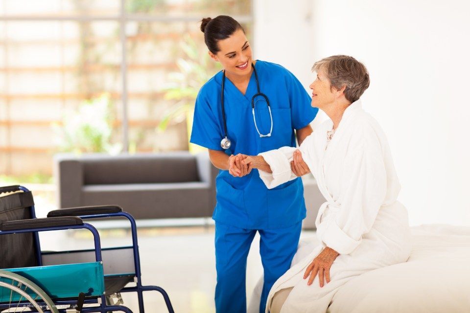 fairpflegt GbR – eine Pflegerin in blauen Scrubs und mit Stethoskop hilft einer älteren Dame vom Bett in den Rollstuhl
