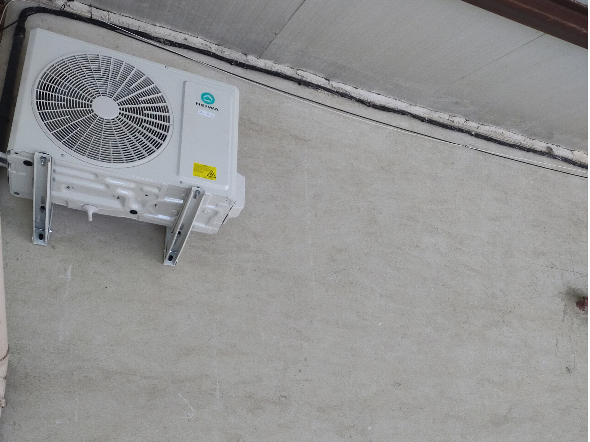 Unité de climatisation extérieure fixée sur un mur en béton gris clair.