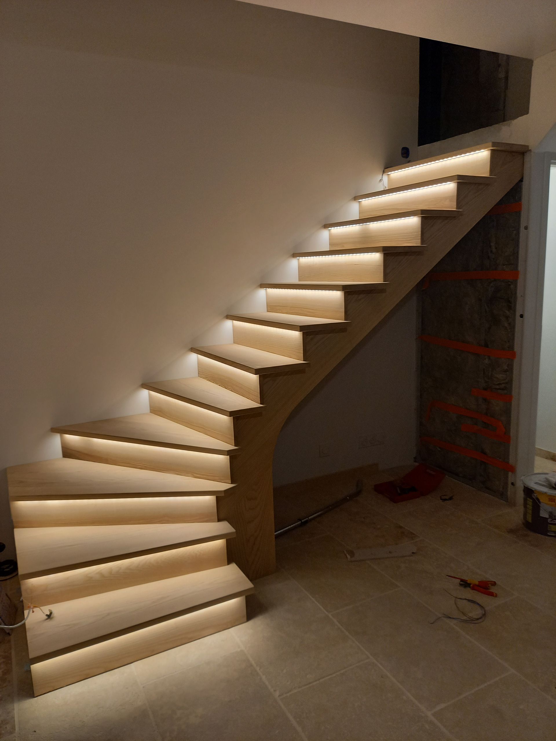 Escalier en bois avec marches éclairées, adossé à un mur blanc et un côté partiellement fini.