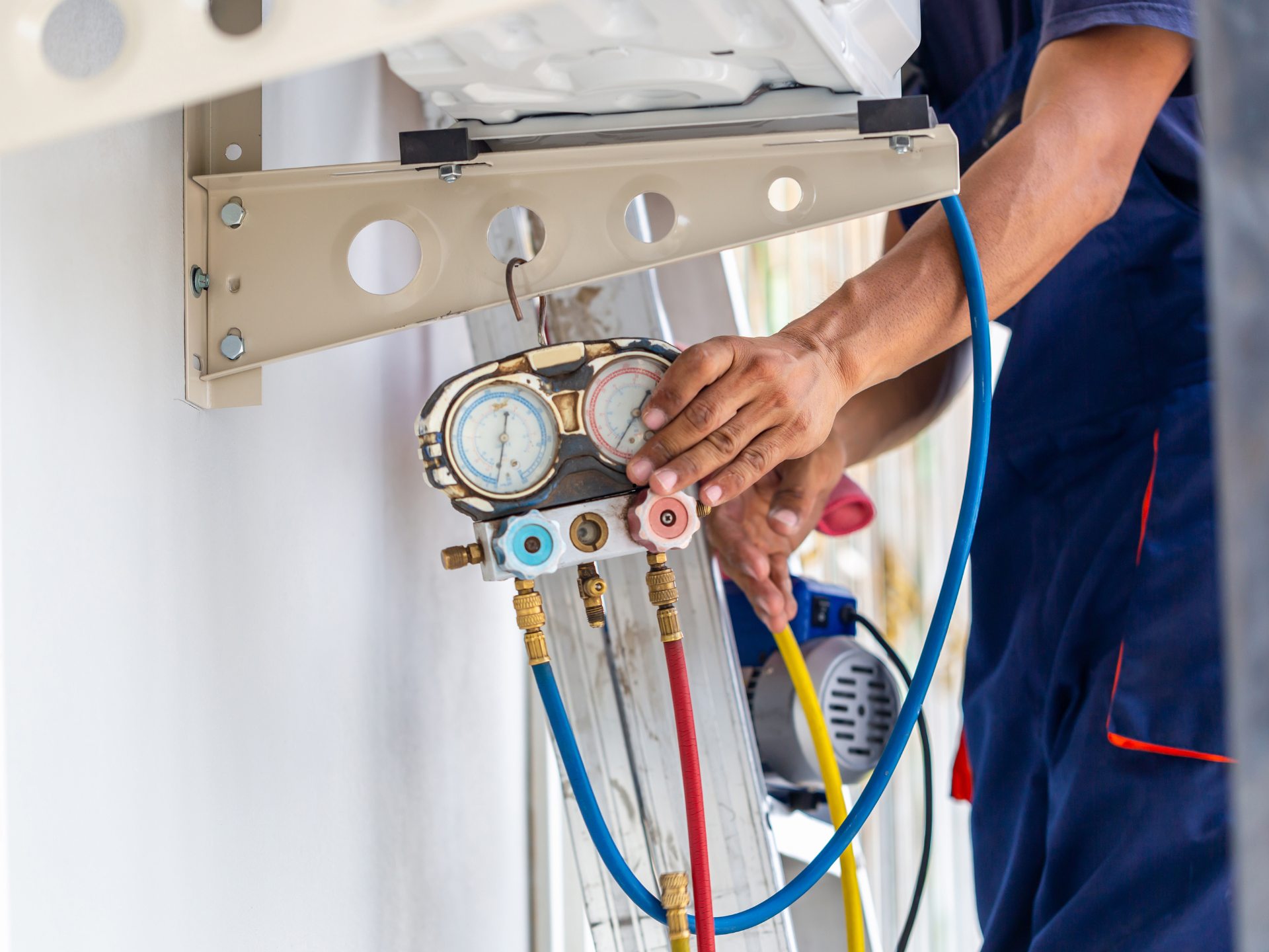 Un technicien en salopette bleue répare un climatiseur. Il règle des manomètres auxquels sont raccordés des tuyaux.