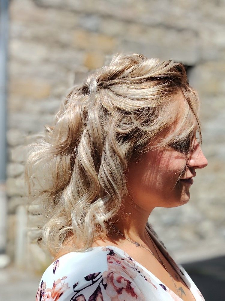 Femme blonde aux cheveux ondulés, portant un haut fleuri, devant un bâtiment en pierre.