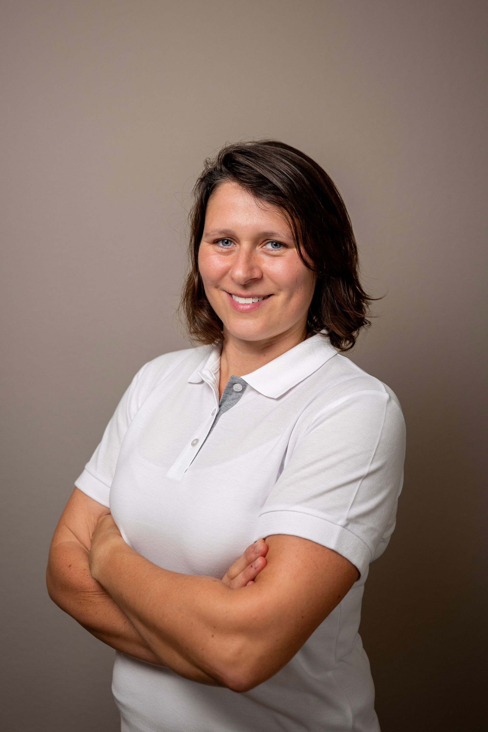 Frau mit verschränkten Armen, lächelnd, in einem weißen Poloshirt, vor einem beigen Hintergrund.