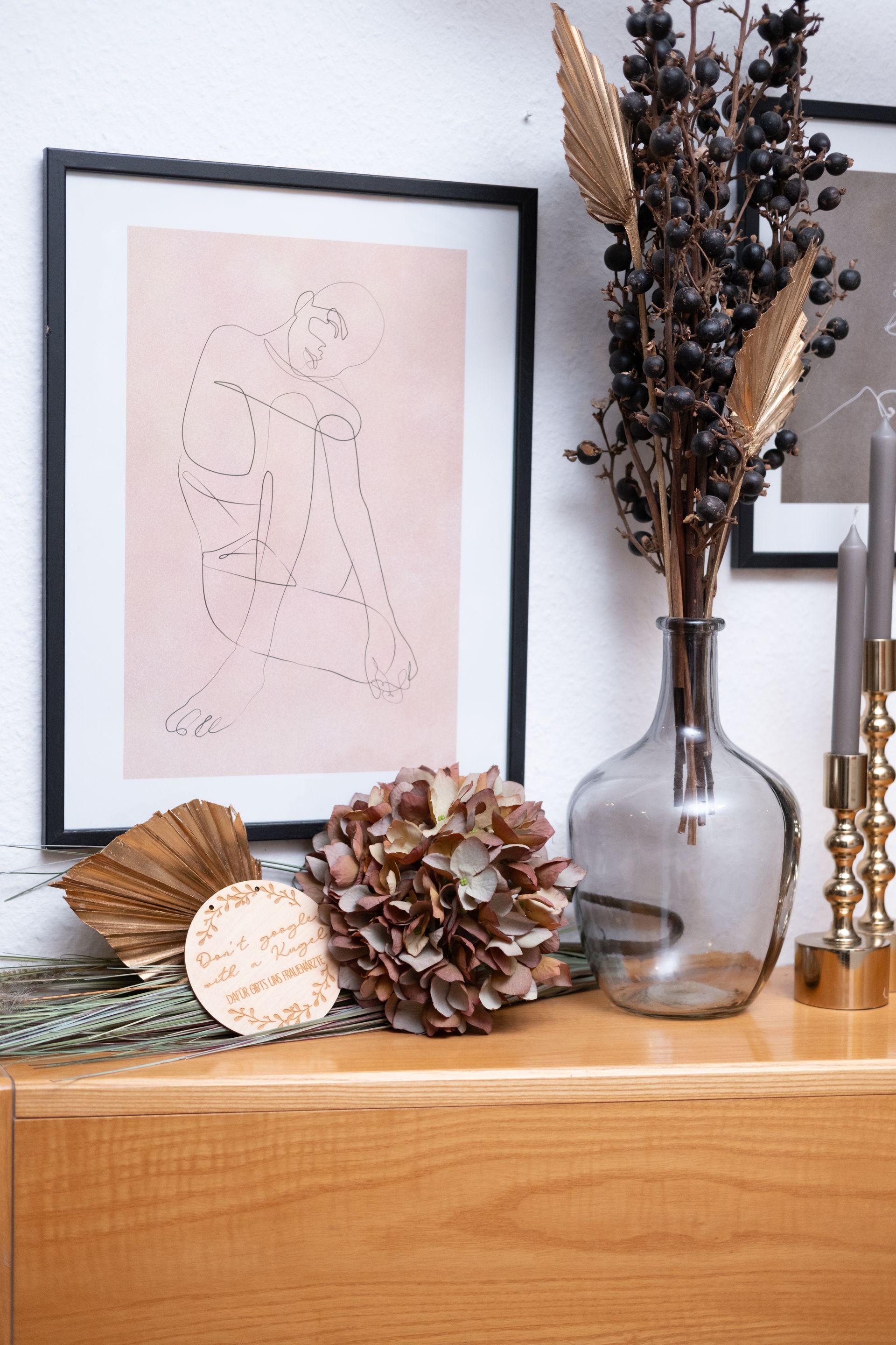 Eine Vase mit getrockneten Blumen steht auf einem Holztisch neben einem gerahmten Bild.