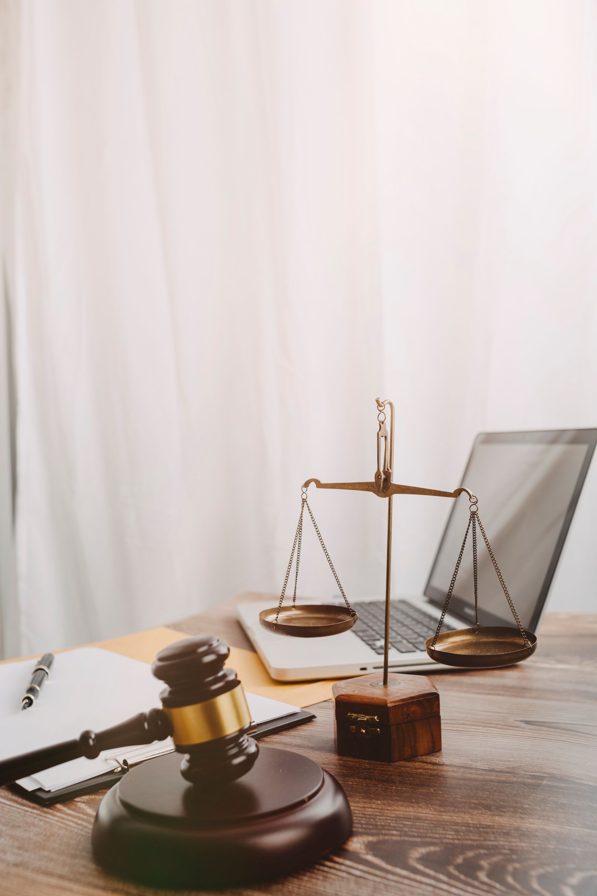 Des livres de droit, un maillet et une balance de la justice sur un bureau, devant un rideau clair.
