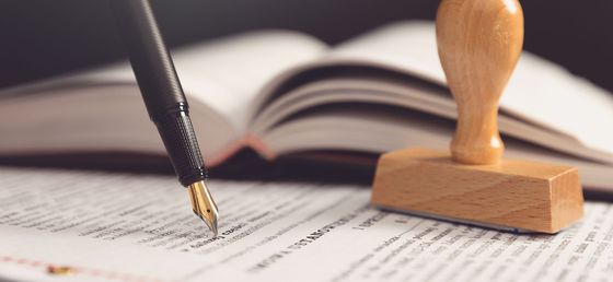 Écriture au stylo sur du papier à côté d'un livre ouvert et d'un tampon en bois sur un bureau.