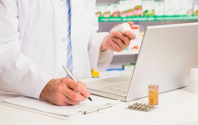 Un farmacéutico sostiene un frasco de pastillas y escribe en un portapapeles frente a una computadora portátil.