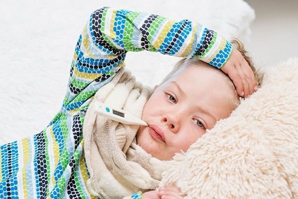 Niño enfermo en la cama, con termómetro en la boca, aspecto cansado, sujetándose la cabeza con la mano.
