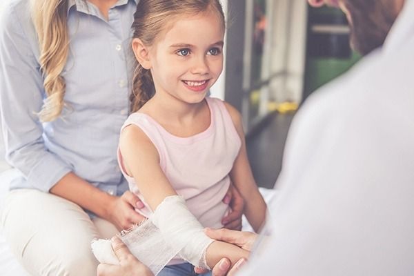 Una niña con un brazo vendado sonríe a un médico y su madre está cerca en un entorno médico.