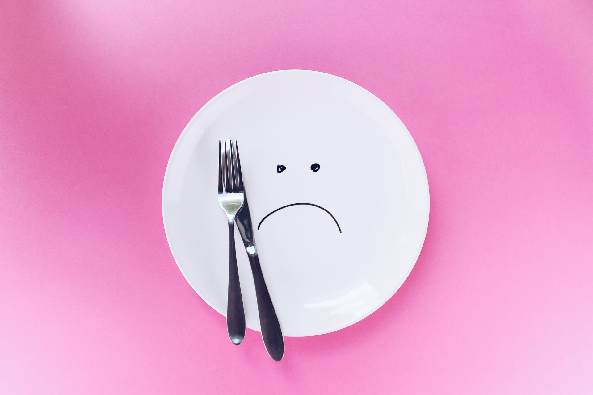 Plato blanco con una cara triste dibujada, tenedor y cuchillo dispuestos como lágrimas, sobre un fondo rosa.