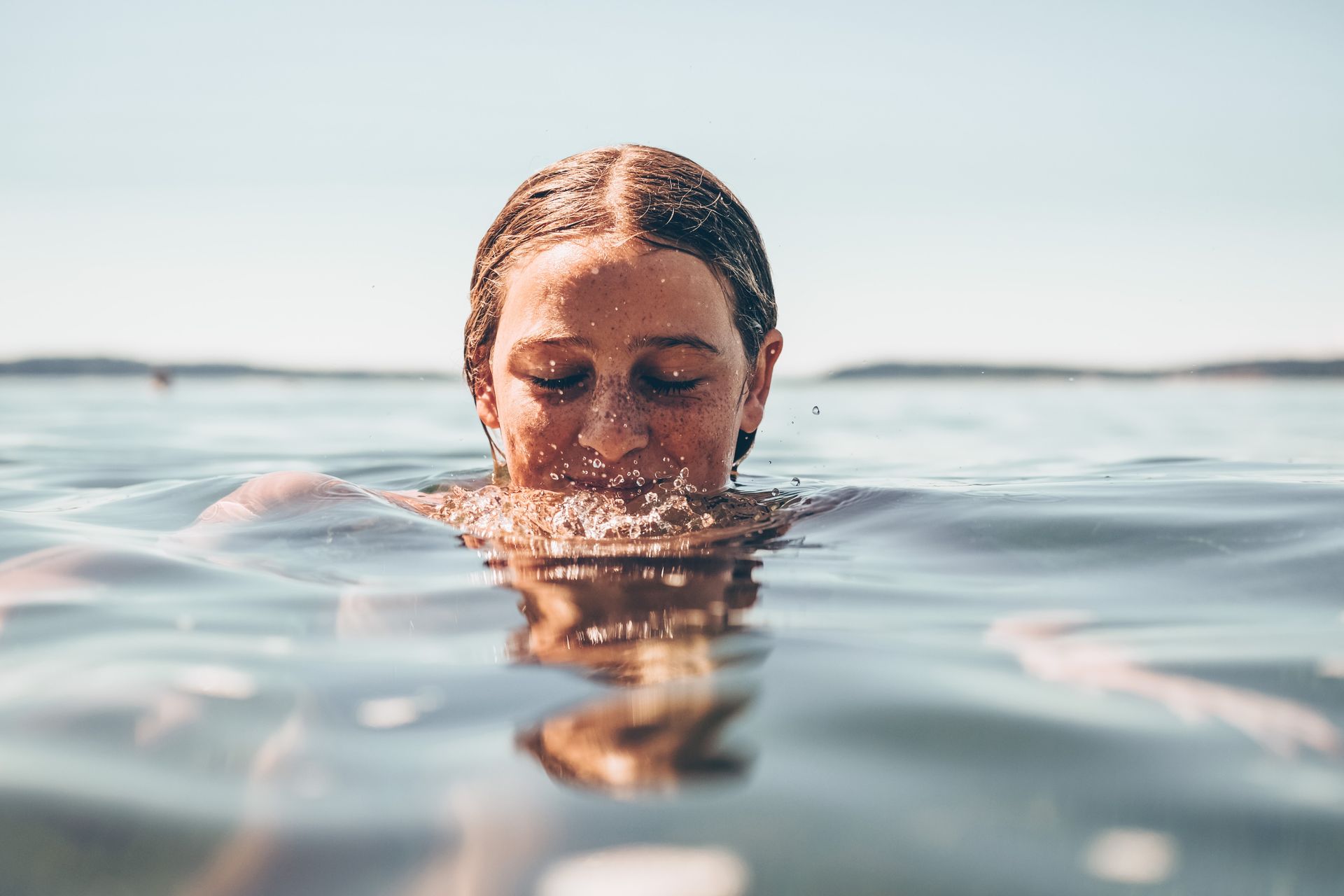 Mujer emergiendo del agua, con los ojos cerrados y gotas salpicando. Cielo azul y océano.