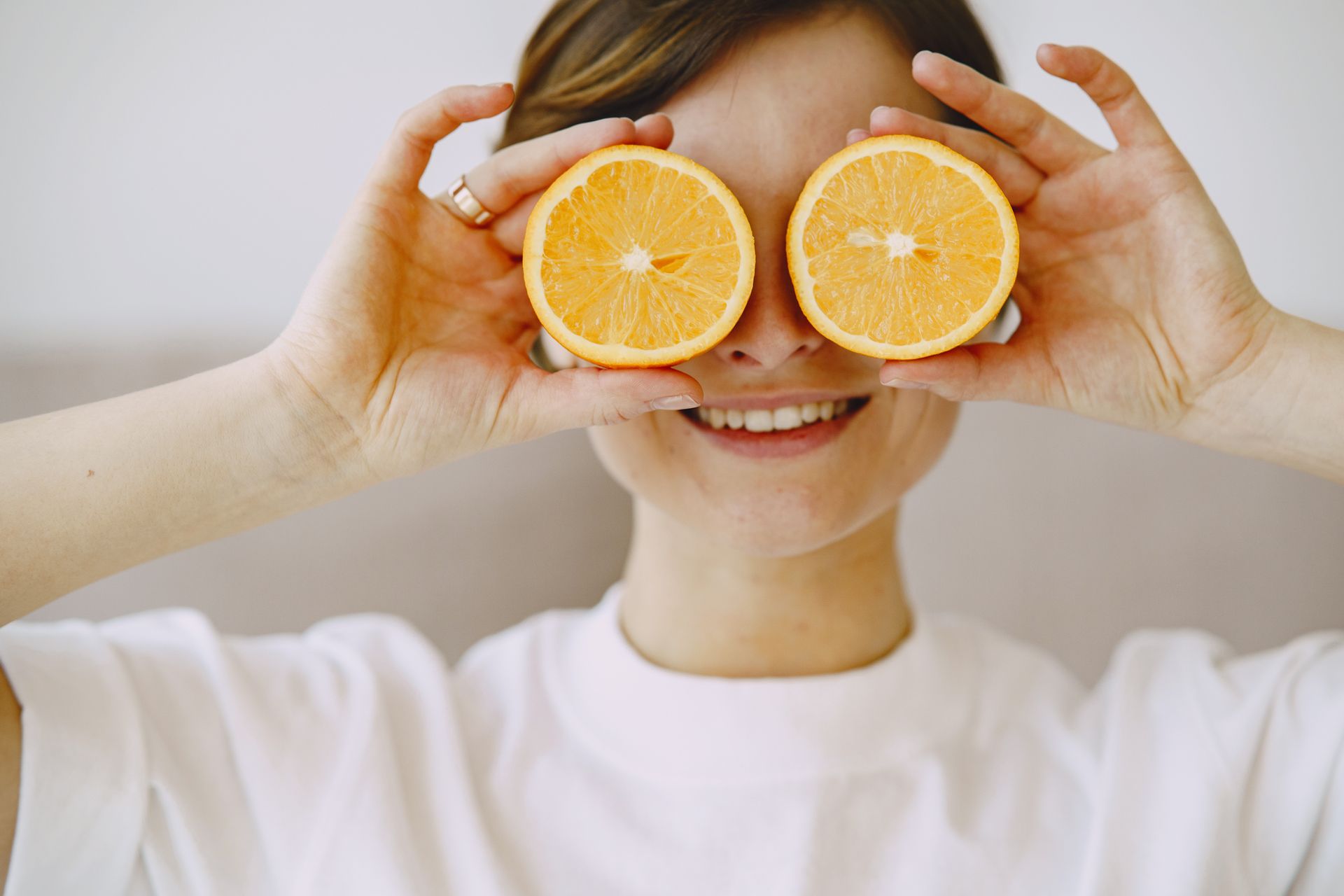 Mujer sosteniendo mitades de naranja sobre los ojos, sonriendo. Camisa blanca, fondo neutro.