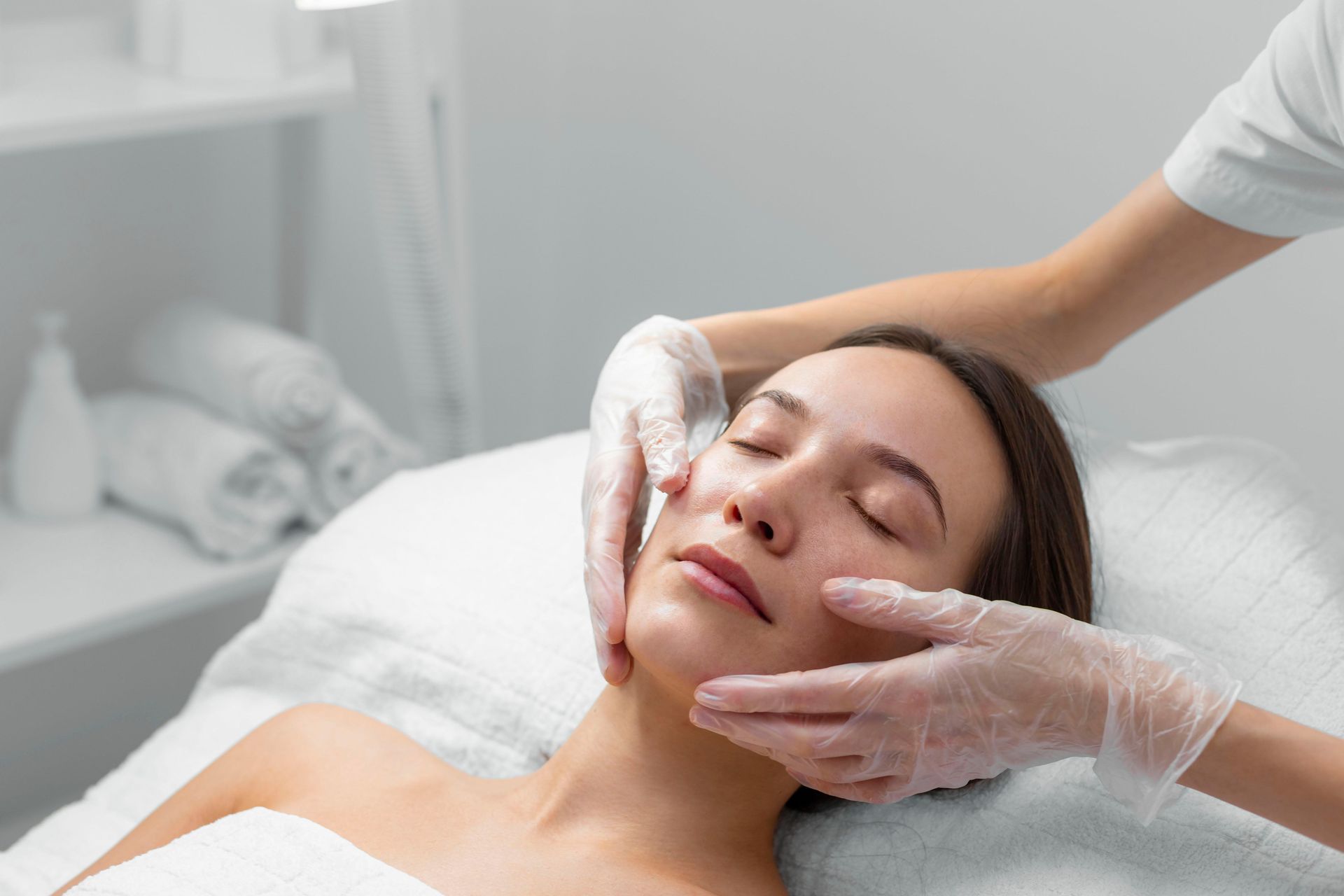 Mujer recibiendo un tratamiento facial con guantes. Está tumbada, con los ojos cerrados, en un spa.