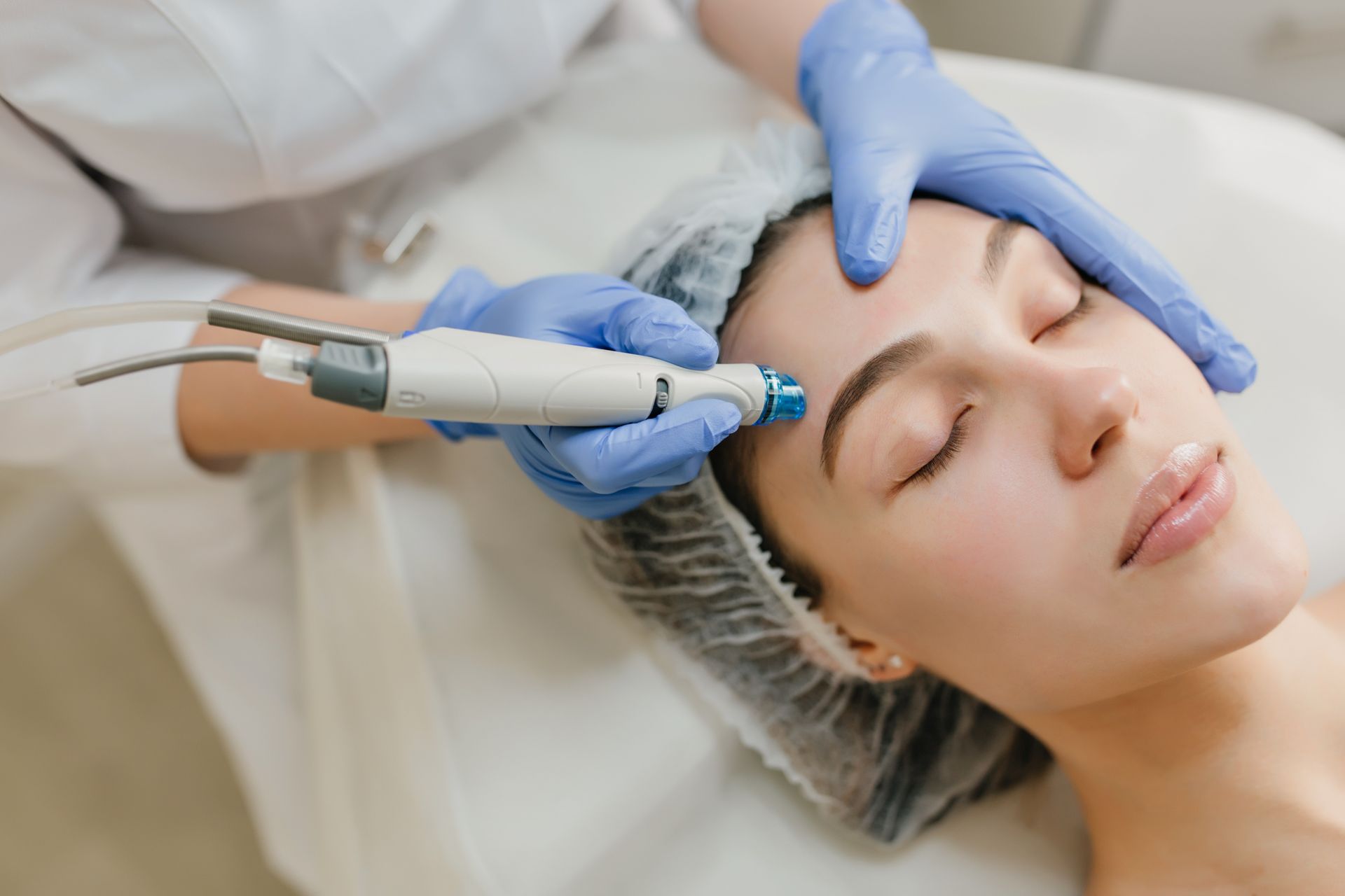 Una mujer recibe un tratamiento facial con guantes azules. Está tumbada en una camilla blanca.
