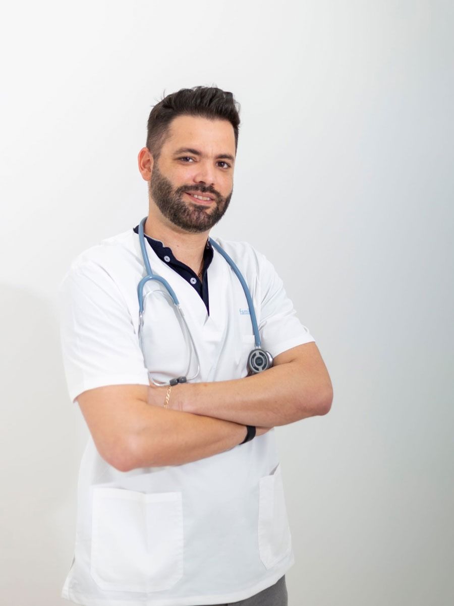 Médico con los brazos cruzados, estetoscopio, sonriendo, vistiendo bata blanca, de pie contra un fondo blanco.
