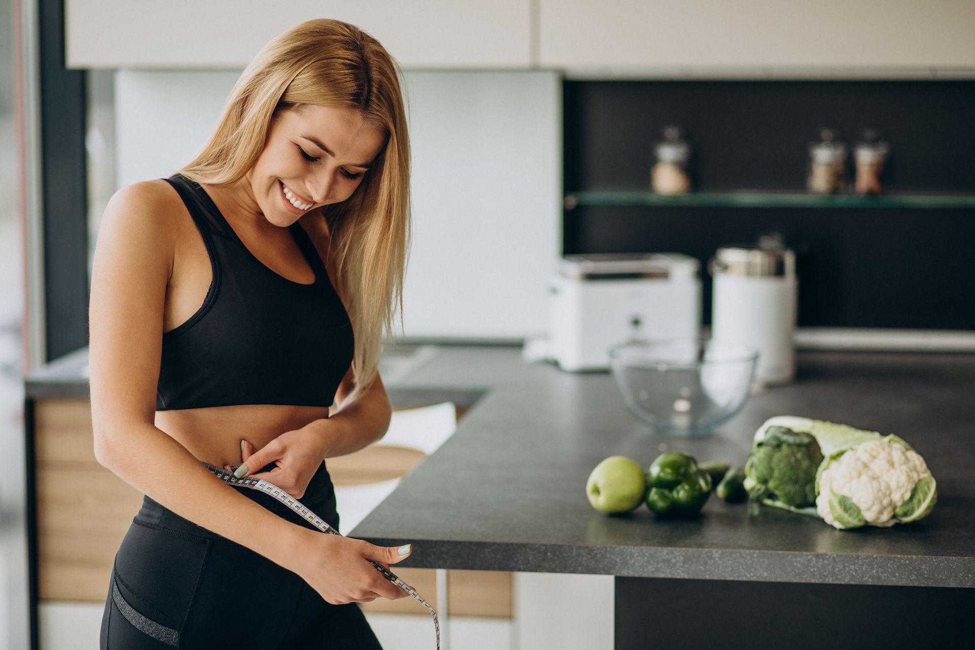 Mujer en ropa deportiva midiendo la cintura en la cocina con verduras.