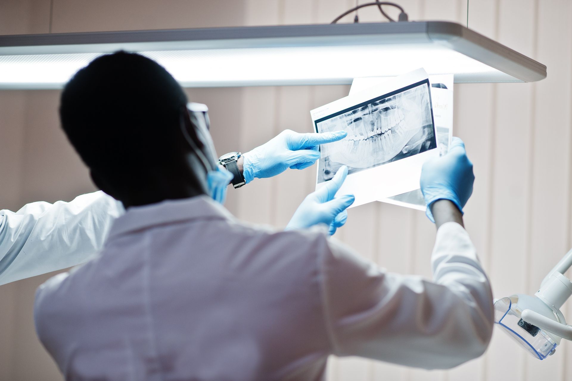 Dos dentistas con batas de laboratorio examinan una radiografía de dientes, uno de ellos señala con una mano enguantada.