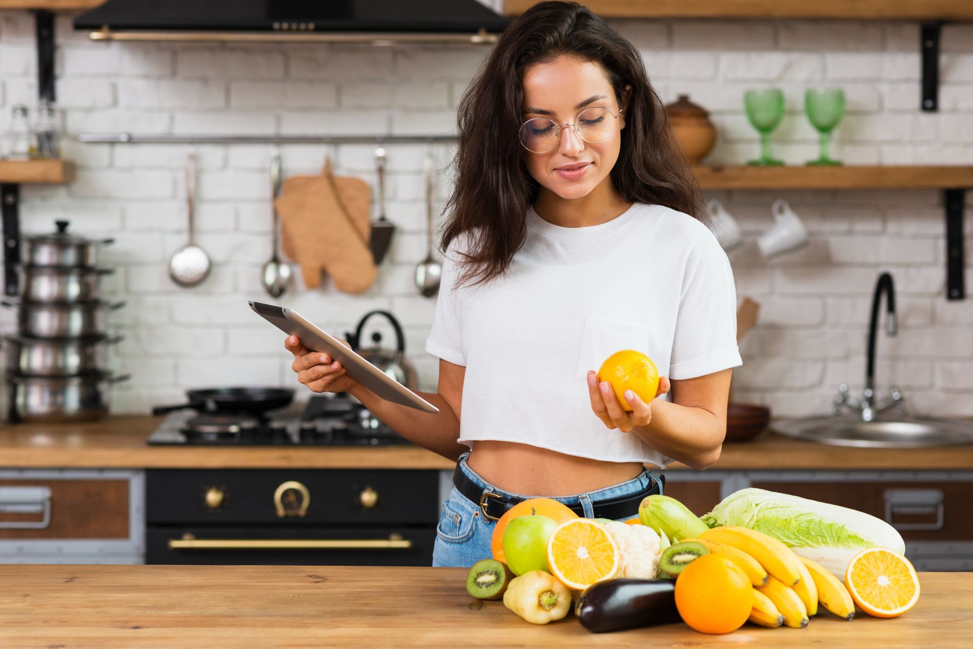Mujer en la cocina sosteniendo una naranja y una tableta, rodeada de frutas y verduras.