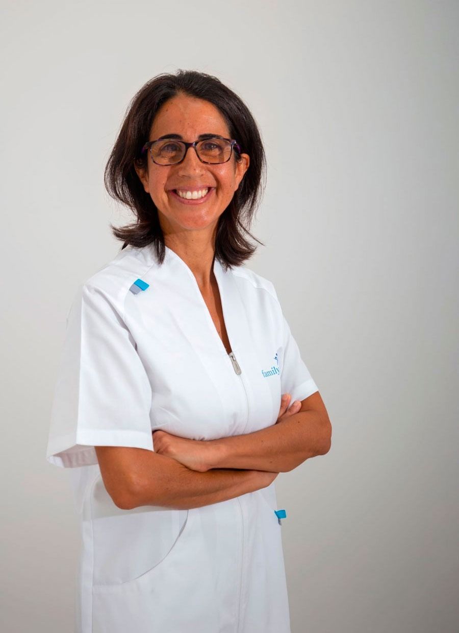 Mujer con uniforme blanco y gafas, sonriendo y con los brazos cruzados, sobre un fondo blanco.