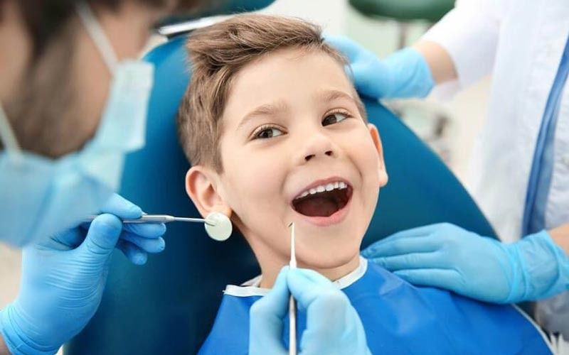 Niño en el dentista, sonriendo mientras le examinan la boca con herramientas, guantes azules y batas.