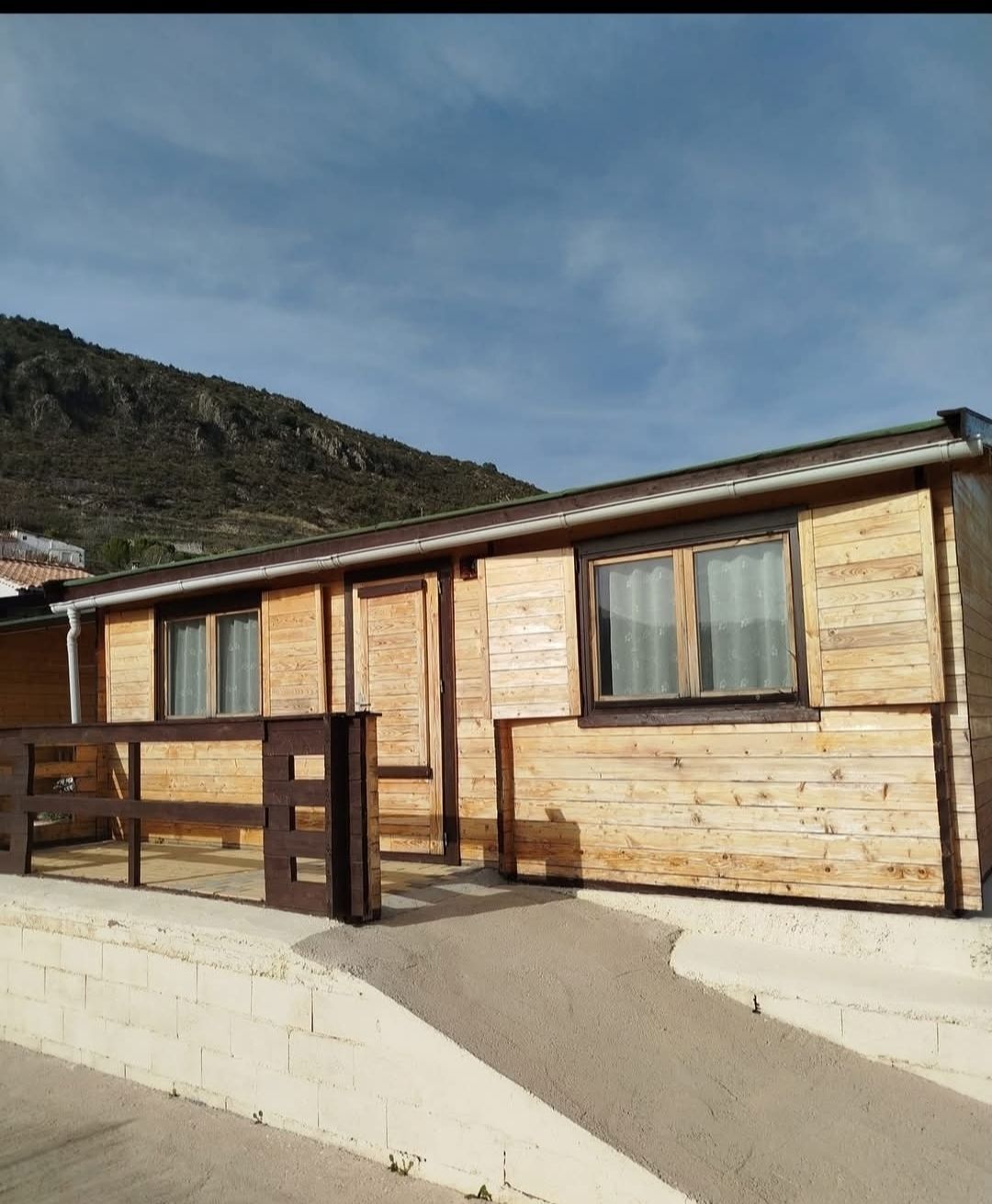 Cabaña de madera con molduras y ventanas marrones, una pequeña valla y un fondo de montaña.